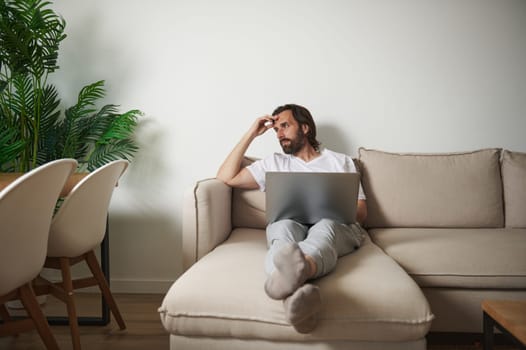 Professional man working remotely on a laptop in a cozy living room space, reflecting on ideas. Surrounded by greenery and furniture, creating a serene telecommuting environment.