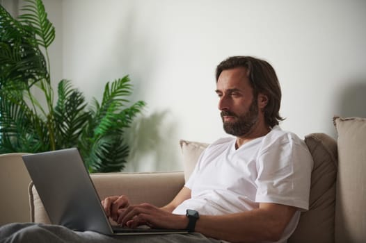 Adult male engaged in online work, typing on a laptop while seated on a sofa in a natural home setting. Green plants add a cozy ambiance.