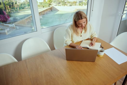 A woman working remotely at a wooden table, utilizing a laptop and notebook. Bright daylight illuminates the modern and cozy workspace, emphasizing productivity and focus.