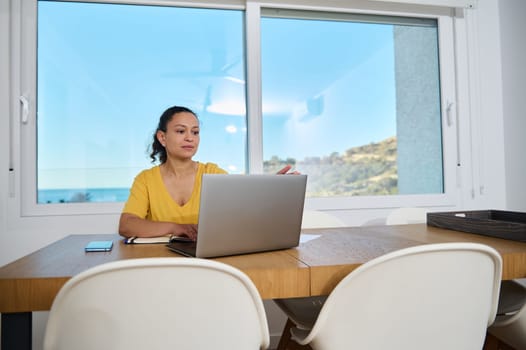 A dedicated professional sits at a wooden table in a serene home office while using a laptop, reflecting focused engagement amidst a cozy and picturesque environment.
