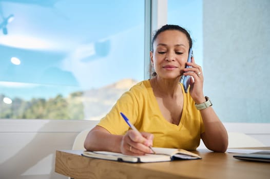 A woman working remotely at her home office, coordinating on a call and writing notes. A modern professional multitasking in a comfortable, bright, and inspiring workspace.