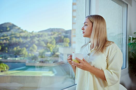 Woman gazes thoughtfully out of a window overlooking a serene landscape, wearing a casual yellow top and holding a mug, radiating calm and introspection.