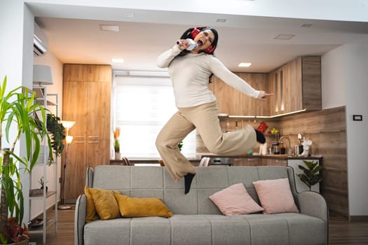 Woman with wireless headphones jumping on sofa and singing at home.