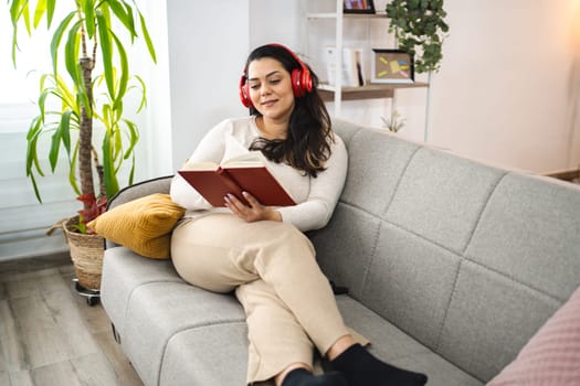 Woman with wireless headphones reading a book on a sofa.