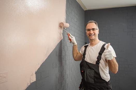A man is seen standing before a wall, methodically applying paint with a roller. He wears work clothes and holds the roller in his hand, focused on the task at hand.
