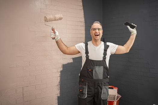 A man wearing overalls is standing while holding a paint roller in his hand, ready to apply paint to a surface.