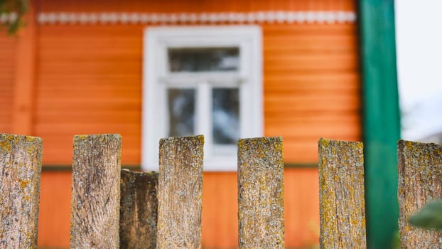 Weathered wooden fence with lichen is standing in front of typical orange russian house with white window