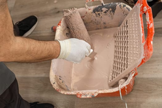 Close up of the container with paint and the paint roller. A man dips a paint brush into the container of paint.