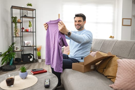 Brown-haired man sits on the couch in the living room and happily opens a cardboard package and looks at a new T-shirt.
