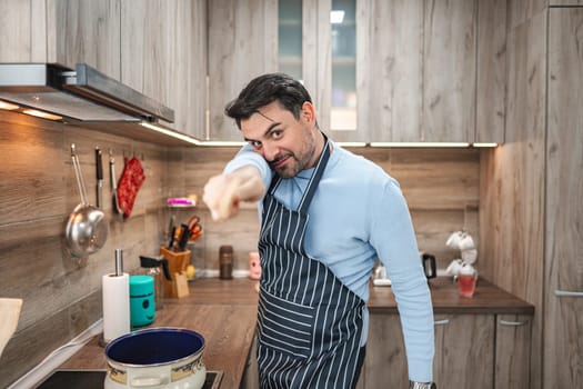 Man with brown hair is standing in front of the stove in the kitchen with a wooden stick in his hand and is having fun while cooking..