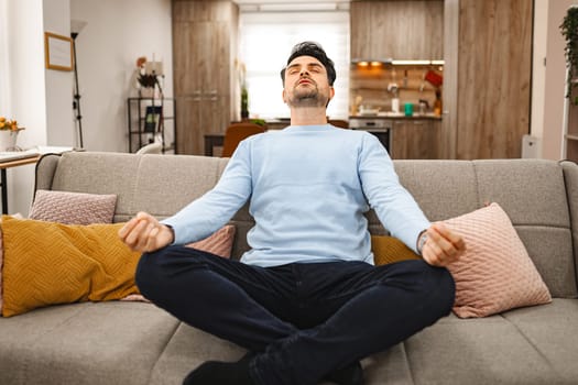 Brown-haired man is sitting relaxed on a couch in a spacious living room, he is meditating.