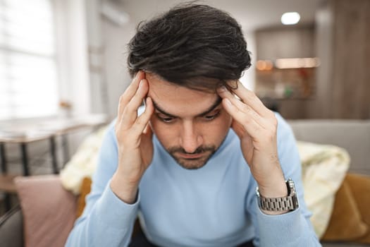 A man is seated on a couch, his hands clasped to his head in evident distress or frustration. He appears deep in thought or experiencing emotional turmoil.