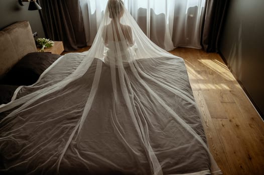 Woman in white dress sitting on bed. She is wearing luxurious white veil. Next to her is bouquet of flowers. View from back in front of white curtain on window in hotel. Silhouette of bride