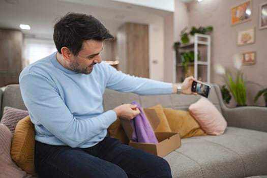 Brown-haired man sitting on the couch in the living room, waving and thanking the interlocutor via video call that the shipment arrived, purple shirt.