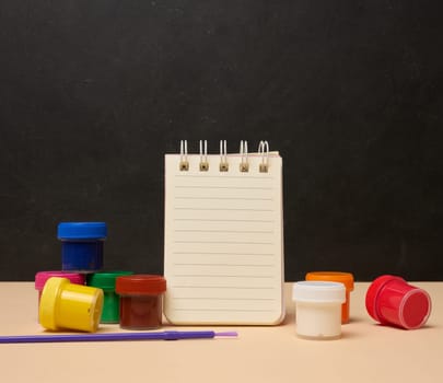 An open white spiral notebook surrounded by colorful paint cans and a brush