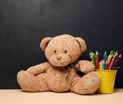 A brown teddy bear sits next to a yellow bucket filled with colorful pencils on a table with a black background.