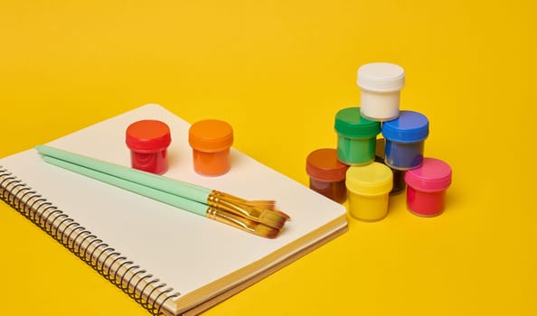 White spiral notebook, brushes and colorful paint cans symbolizing art, creativity or hobby placed on yellow background