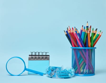 Set of office supplies including colored pencils in a glass, magnifying glass, notepad and paper clips symbolizing learning, organization or creativity, blue background