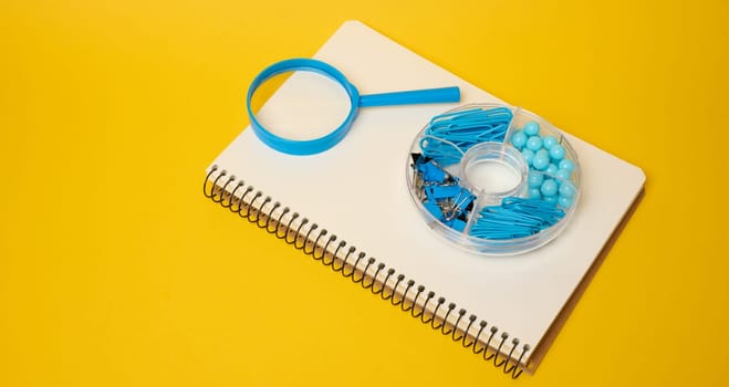 White notebook with blue magnifying glass and blue stationery in organizer symbolizing studying, organizing or creativity placed on yellow background.