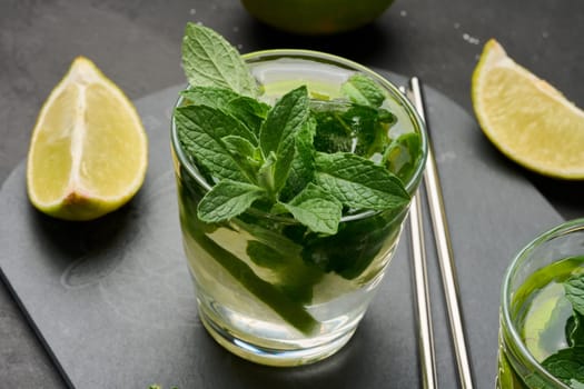 Glass of mojito cocktail with fresh mint leaves, lime slices, and ice cubes on a dark stone background. Refreshing summer drink with stainless steel straws