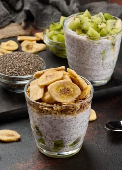 Two glasses of chia seed pudding, one topped with diced kiwi and the other with granola and banana chips, are on a dark surface with ingredients