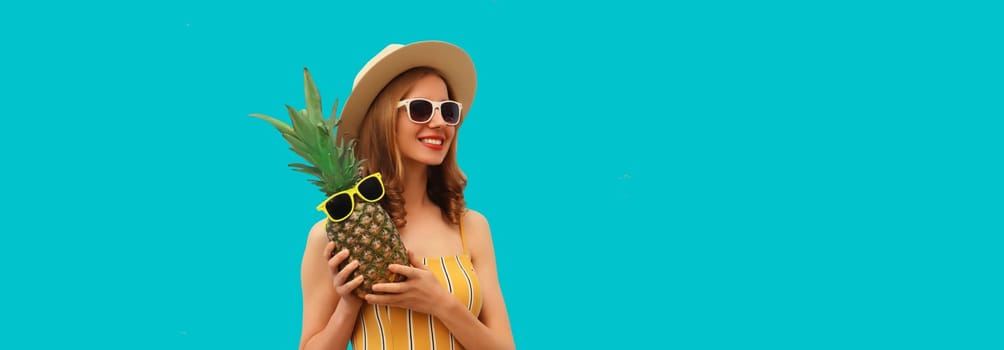 Summer vacation, happy joyful young woman with pineapple fruit in tourist hat, cheerful girl posing on blue studio background