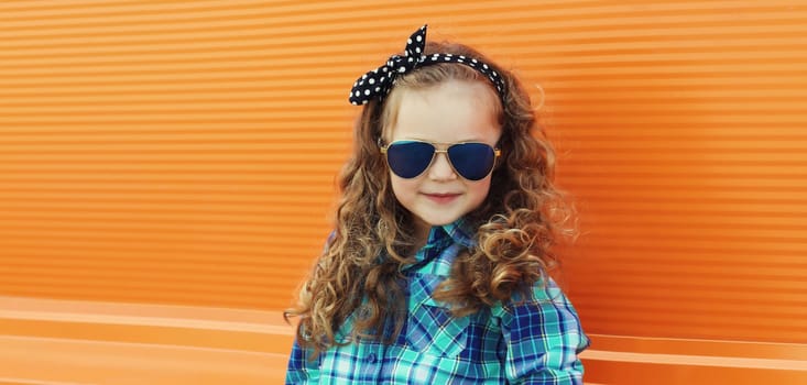 Happy kid girl with curly hair, stylish little child posing on city street on orange wall background