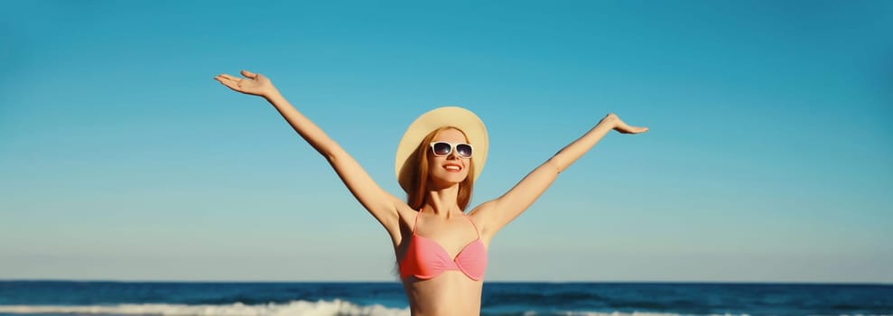 Summer vacation, beautiful happy slim woman in bikini swimsuit and tourist straw hat raising her hands up on the beach on sunny day on sea coast