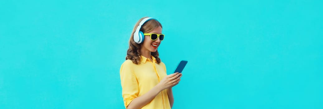 Summer portrait of modern happy young woman listening to music with smartphone, girl smiles in headphones holding phone on bright blue background