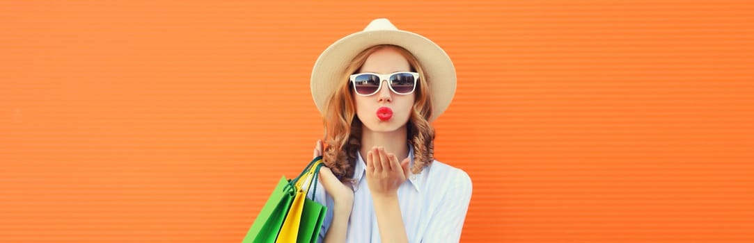 Stylish beautiful young woman model with shopping bags blowing a kiss in summer straw hat on orange studio background