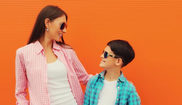 Stylish happy smiling mother with son teenager together in casual clothing in the city on orange wall background, looking at each other