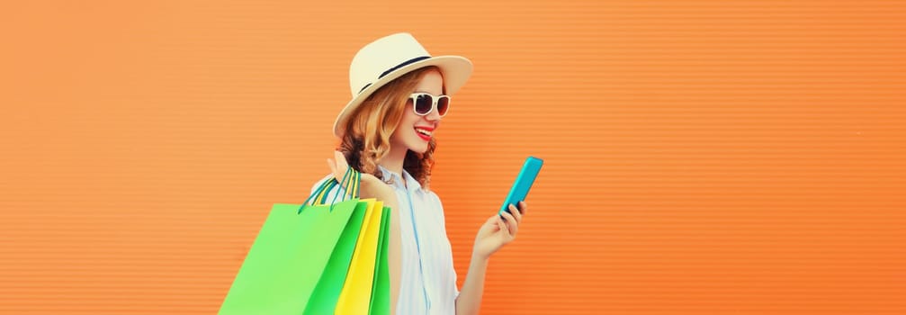 Beautiful happy young woman looking at mobile phone with shopping bag, wearing summer hat on orange studio background