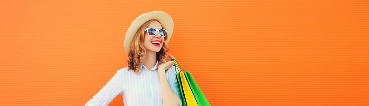 Fashion, purchase. Portrait of stylish beautiful happy young woman posing with colorful shopping bags, girl in summer hat standing on bright orange background