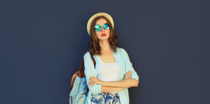 Portrait of stylish modern young woman in summer tourist hat with backpack, glasses and shorts posing on black wall background