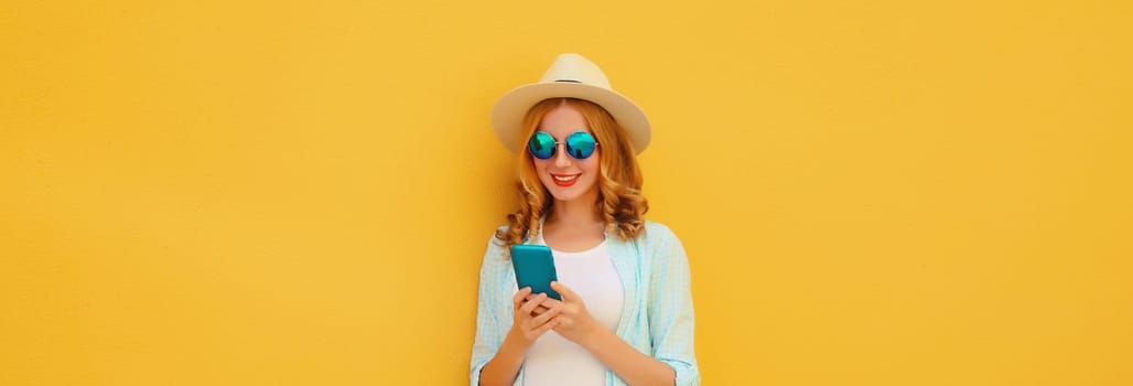 Summer portrait of happy traveler young woman with mobile phone looking at smartphone wearing a straw tourist hat, casual clothing on yellow studio background