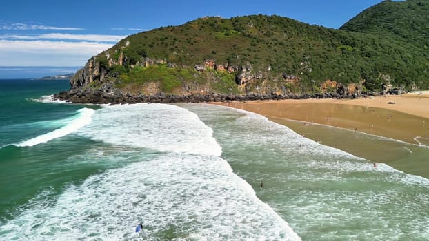 An aerial shot of a peaceful beach featuring gentle, rolling waves, lush green hills, and a clear, bright blue sky