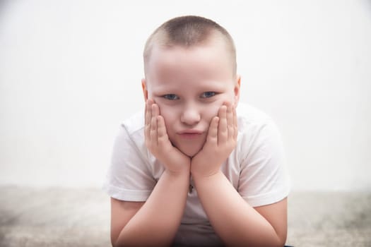 Young boy with short hair shows a pouting expression while resting his hands on his cheeks in a minimalist environment.