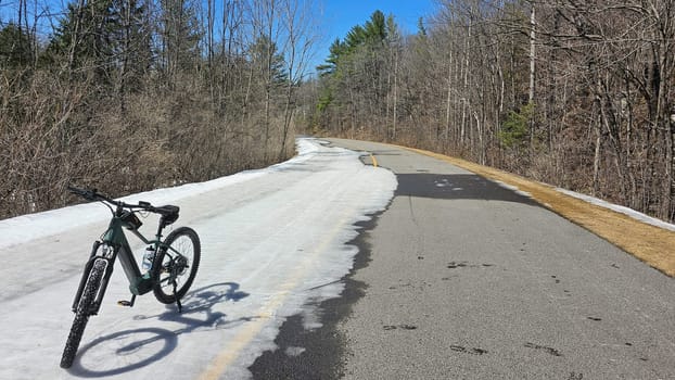 A bicycle rests on a snow-covered road as winter transitions to spring. The scene features bare trees under a clear blue sky, suggesting outdoor recreation and nature exploration.