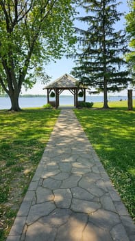 A picturesque stone pathway leads to a beautiful wooden gazebo beside a tranquil lake, surrounded by vibrant green grass and tall, leafy trees, creating a serene atmosphere on a sunny summer day.