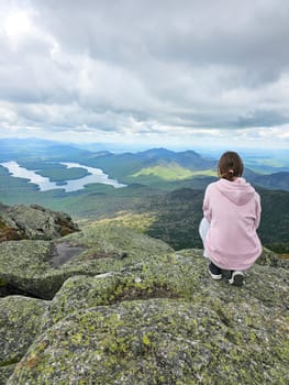 A young girl stands atop a mountain, gazing at the stunning view of Lake Placid and the Adirondack Mountains. This image captures the essence of adventure and the beauty of nature.