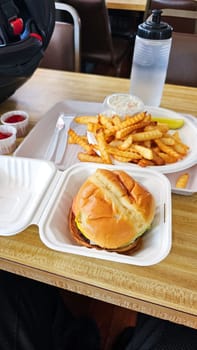 A delicious burger in a clamshell container with crinkle-cut fries on a plate, condiments nearby, and a refreshing drink at a diner, perfect for a lunch after motorcycle riding.