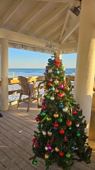 A vibrant array of baubles decorates a festive Christmas tree on a beachfront deck, with the ocean as a backdrop, creating a cheerful holiday ambiance for Christmas celebrations.