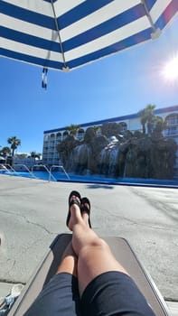 A man lounges by the pool under a blue and white umbrella. He wears flip flops and shorts. A waterfall and resort building are in the background, creating a perfect vacation vibe.