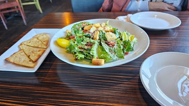 A fresh Caesar salad is beautifully presented on a dark wood table, accompanied by crispbread and empty plates, inviting a delightful dining experience focused on healthy eating.