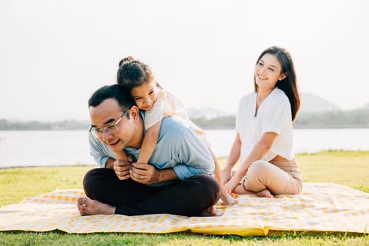 Asian family sitting playing on picnic blanket spring meadow field grass in park, Happy parents child girl riding father back enjoying outdoors together, father carrying daughter, Family relaxation