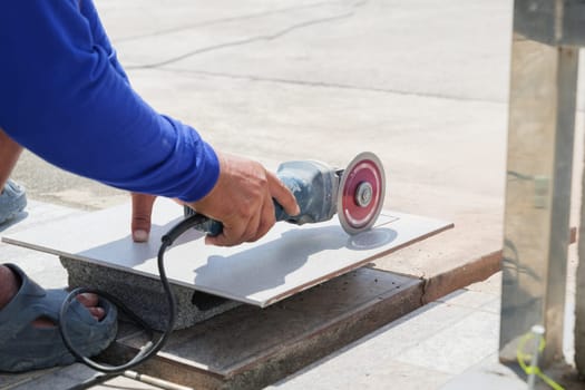 Angle grinder in action: a worker in blue attire carefully cuts a gray tile on a stone surface, showcasing expert tile cutting techniques in construction.