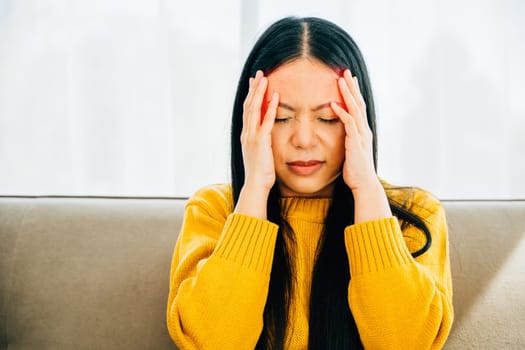 Seated on a sofa an Asian woman manifests stress and headache touching her face. Illustrating health care and medical concept revealing discomfort and exhaustion.
