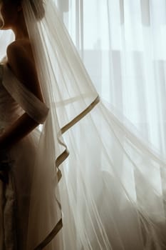 Silhouette of a blonde bride with a lush veil, in a long white dress near a window with a bouquet of lilies. Minimalistic interior, hotel or house. Bride in a long dress
