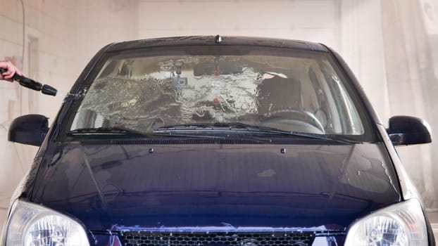Car wash worker using high pressure water jet to clean windshield of dark blue car
