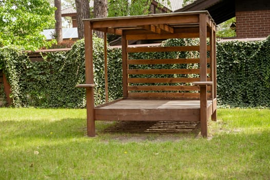 Wooden brown gazebo with a canopy in the middle of the garden. Picnic gazebo among trees, surrounded by a fence with ivy, on a bright lawn.